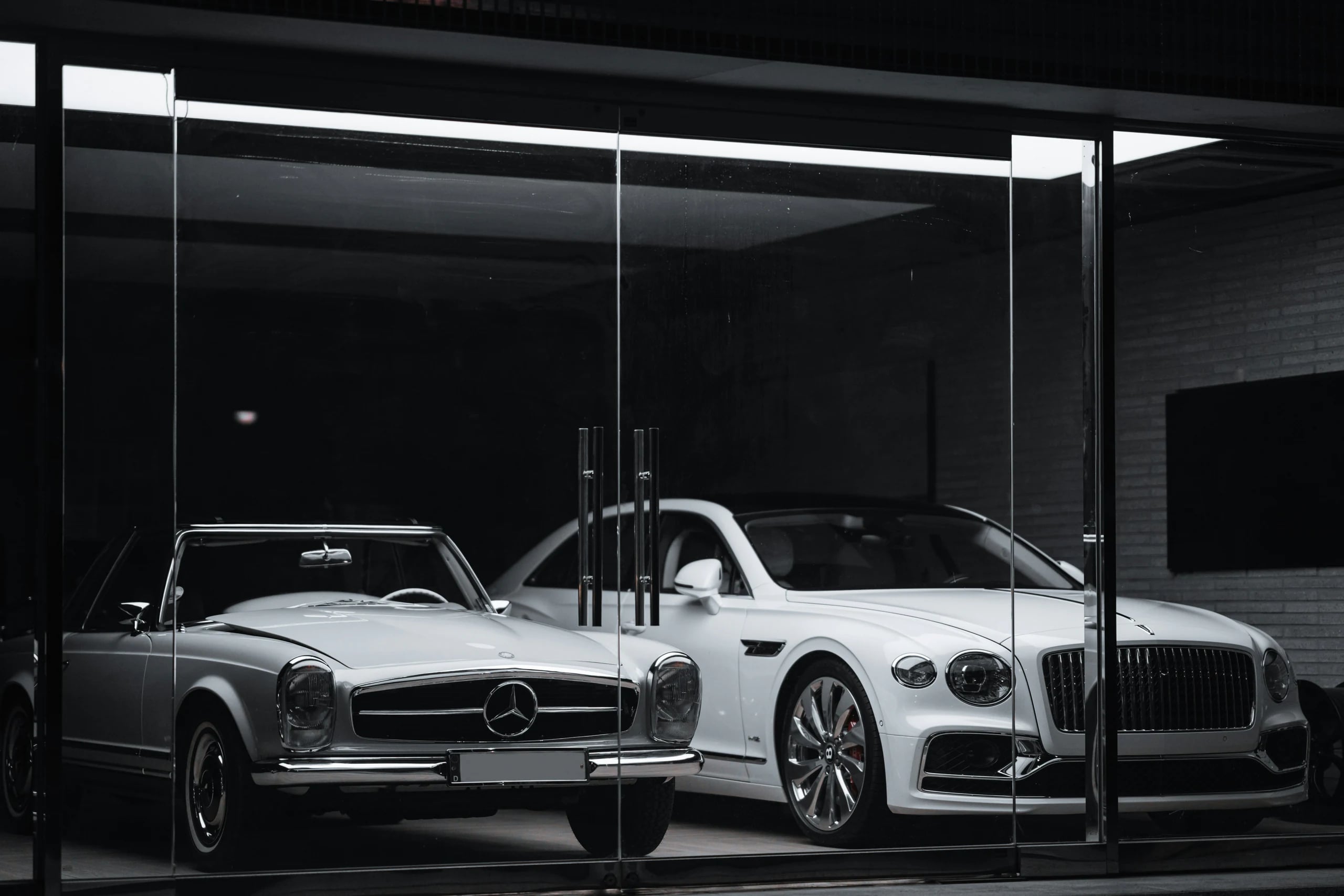 Mercedes classique gris argenté et Bentley blanche exposées derrière la vitrine d'un showroom, le soir